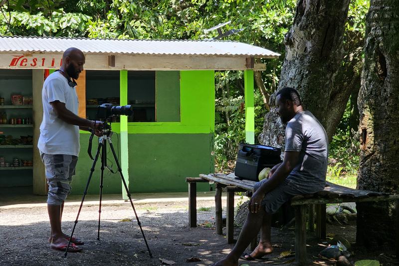 Camera operator filming a man seated at an outdoor bench, with the camera on a tripod