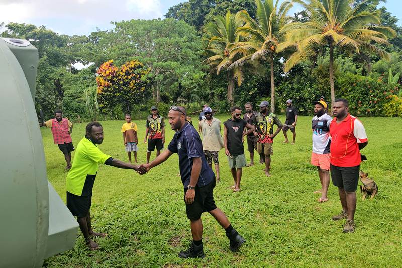 Two men shaking hands beside a large water tank, with a community group on the lawn behind