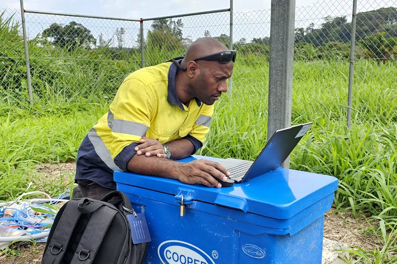 Technician in hi-vis using a laptop on a cool box beside a fenced station